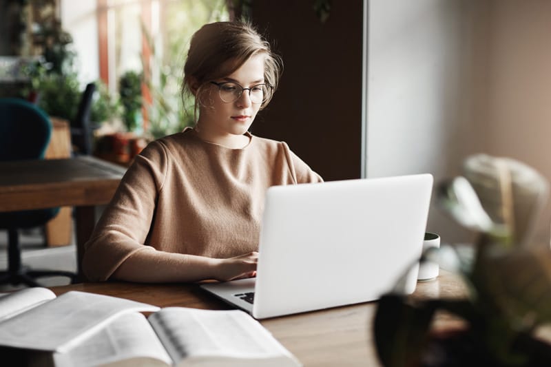 work lifestyle and business concept good looking focused european female in trendy glasses sitting in cafe near laptop working on notebook surrounded with books making notesjpg